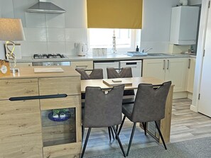 A modern kitchen with a dining area, featuring a wooden cabinet, a built-in stove, and a window with a view. - 3 bedroom accommodation in Bude (Bude)