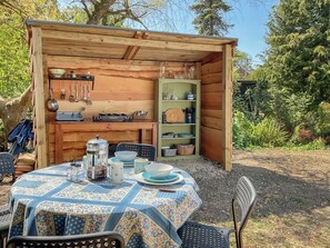 A wooden outdoor dining area with a table set for two, surrounded by a wooden structure with shelves containing kitchenware. - 1 bedroom accommodation in Dorset (Dorset)