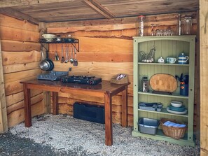 L’intérieur d’un chalet en bois comprend une table en bois, une cuisinière à gaz, une étagère avec des ustensiles de cuisine et un buffet vert contenant de la vaisselle et des ustensiles de cuisine.