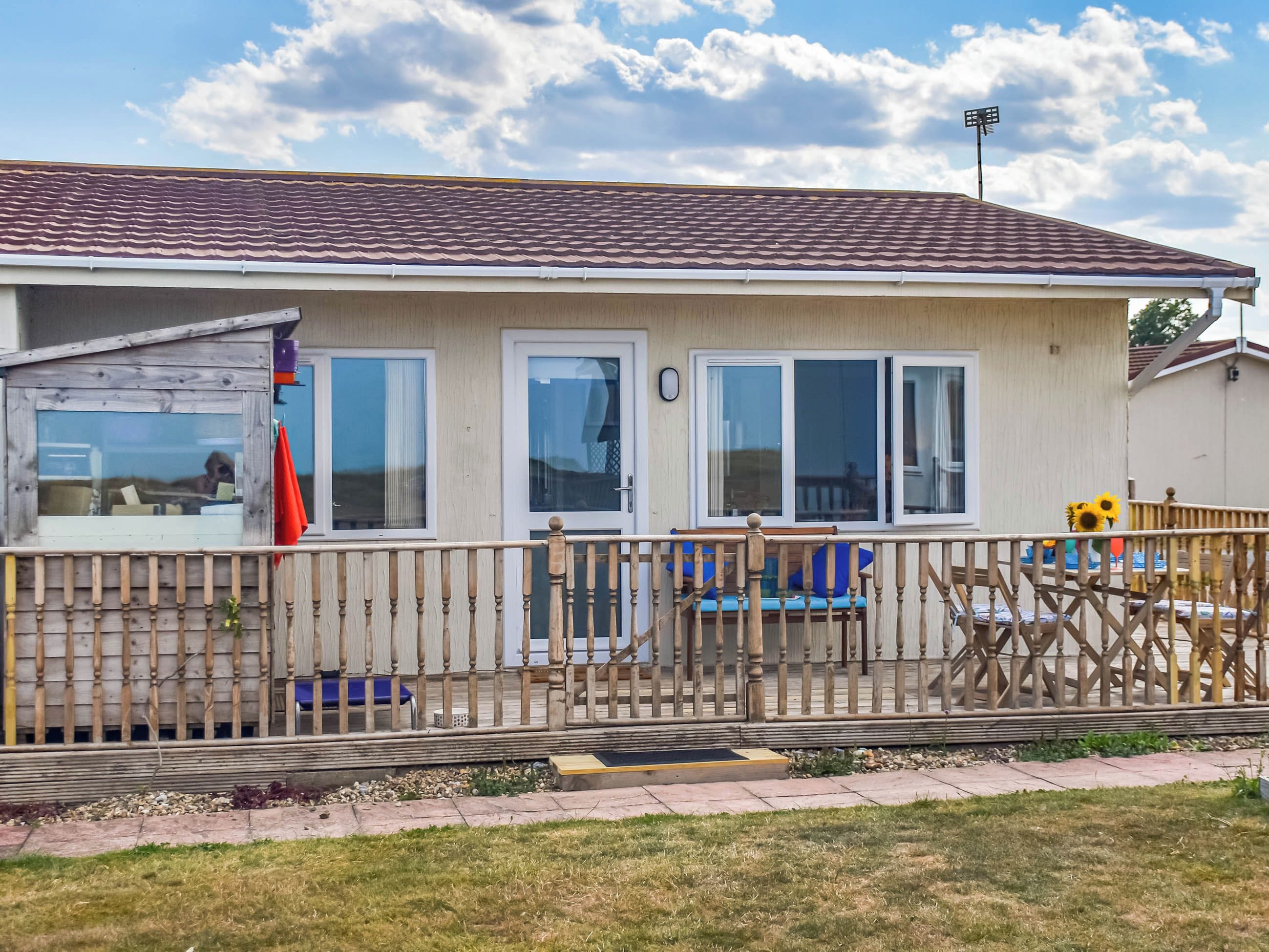 A single-story house with a tiled roof, a wooden deck, and a fenced outdoor area with a bench and a table.