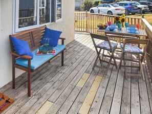 A wooden deck with a bench and chairs, a table with a bowl of fruit and glasses, and a view of parked cars in the background. - 2 bedroom accommodation in Bacton (Bacton)