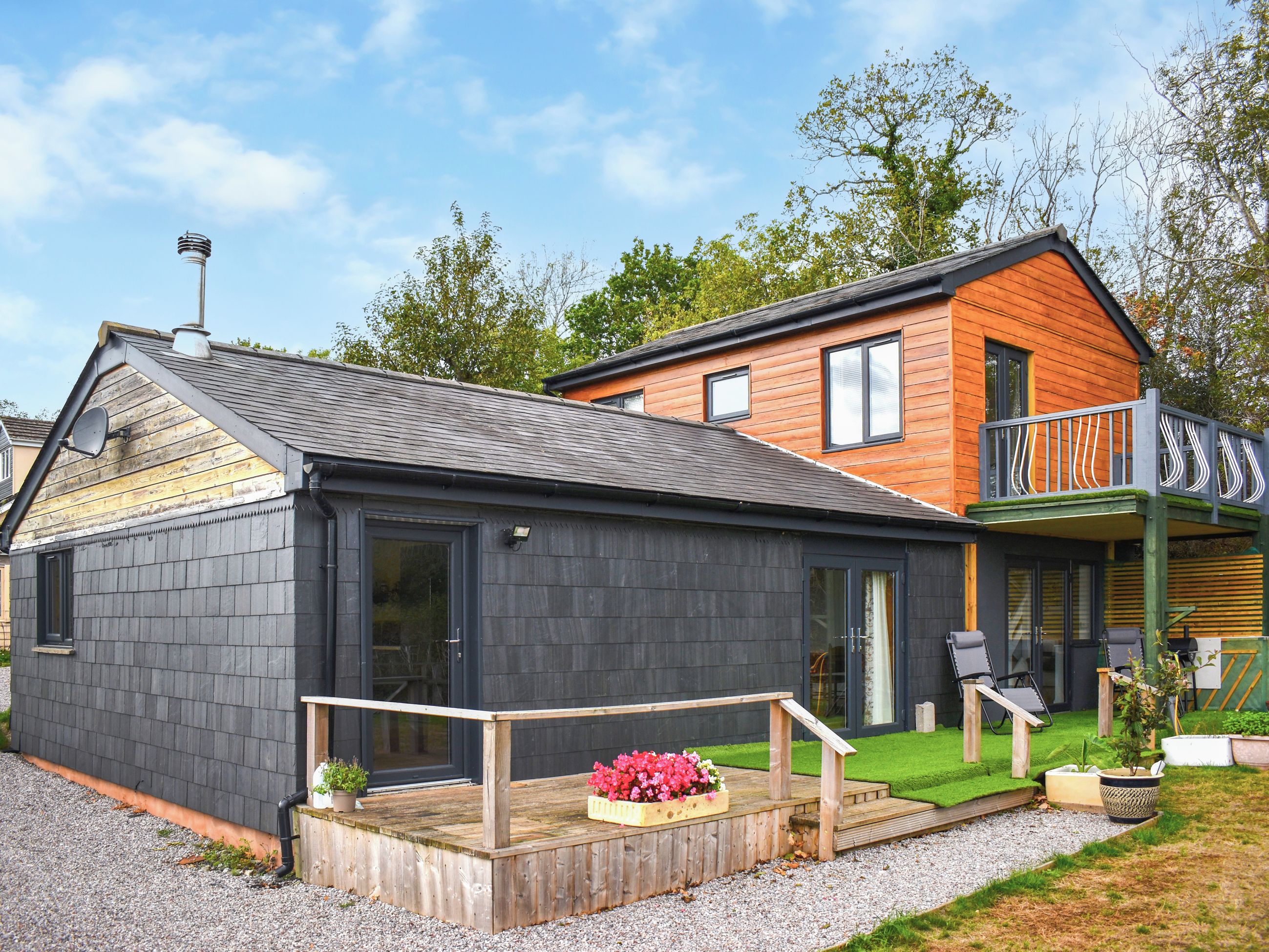 Une maison à deux étages, avec une façade de couleur gris foncé, une terrasse en bois avec des marches et un petit jardin agrémenté de plantes en pot.