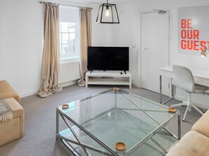 Ett modernt vardagsrum med ett soffbord i glas, en platt-TV, ett vitt skrivbord och en stol.