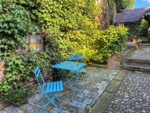 Ein Innenhof mit einem blauen Metalltisch und -stühlen, umgeben von üppiger Vegetation und einem Kopfsteinpflasterweg.