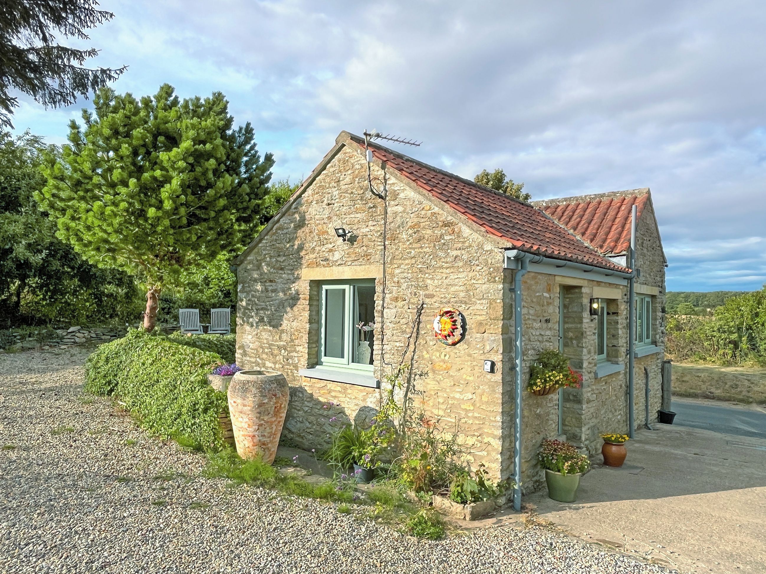 A stone house with a red-tiled roof, a small porch with potted plants, and a gravel driveway.