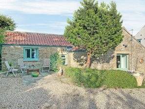 A stone house with a tiled roof, a patio with chairs, and a large tree.