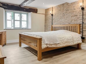 A bedroom with a wooden bed, a stone wall, a window with curtains, and a nightstand. - Lower Tower Hill Farm (Up Holland, West Lancashire)