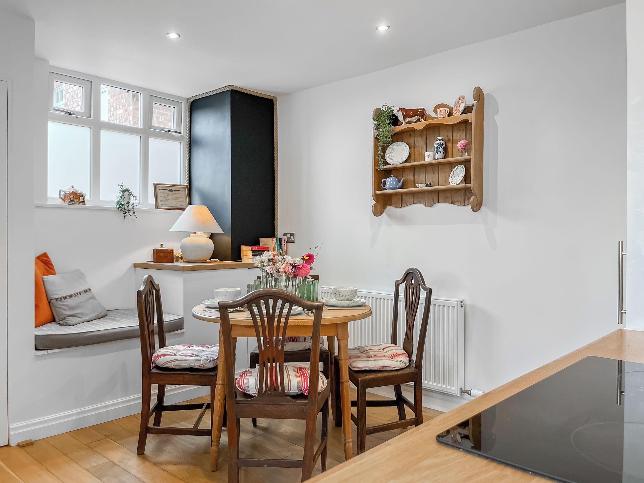 A cozy dining area with a round table, wooden chairs, a small shelf with decorative items, and a window with a view of buildings.