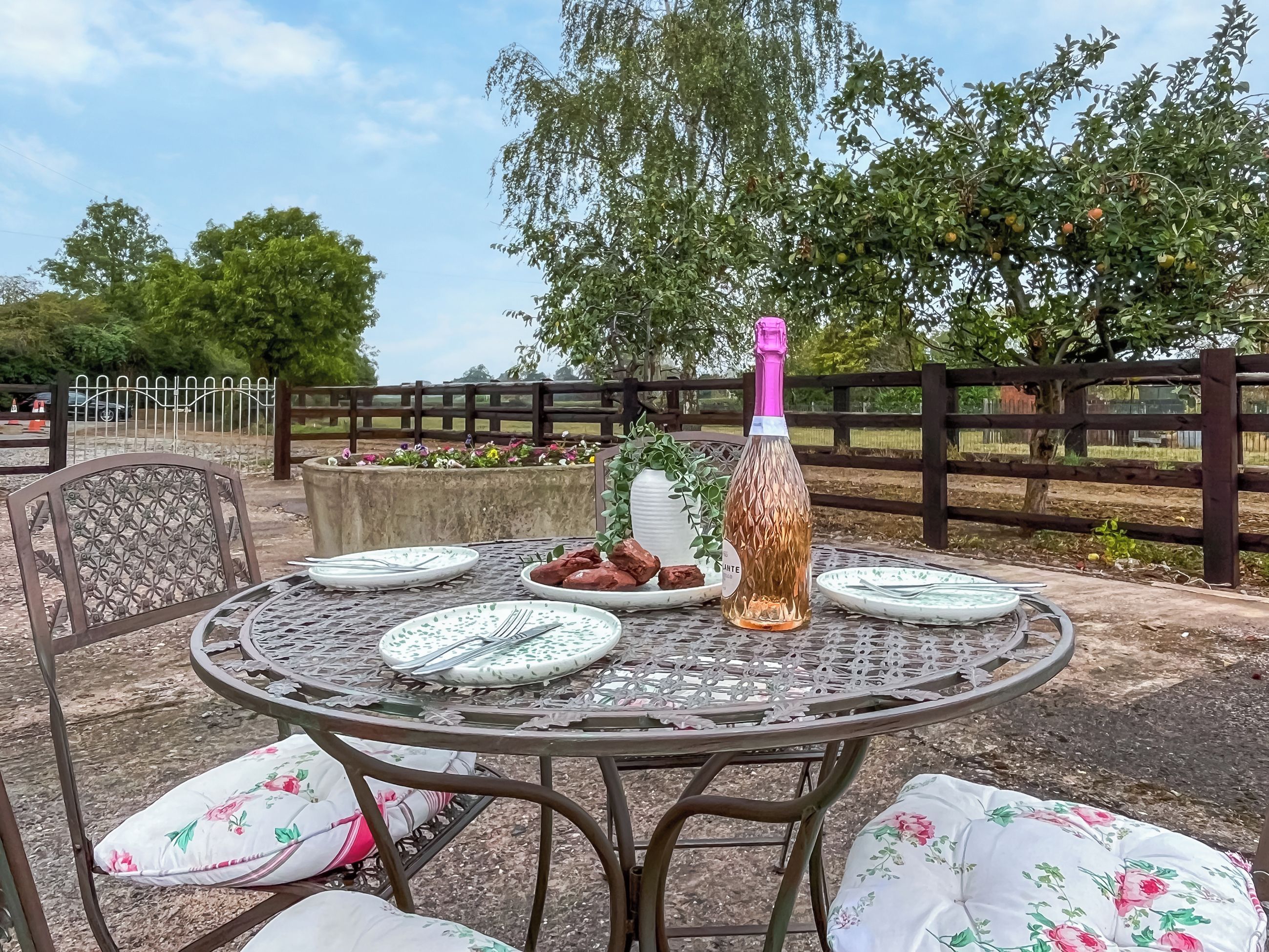 An outdoor dining area with a round metal table, white floral cushions, and a bottle of champagne.