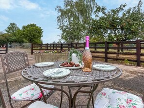 An outdoor dining area with a round metal table, white floral cushions, and a bottle of champagne. - Edleston Farm Cottage - UK52358 (Edleston, near Nantwich)