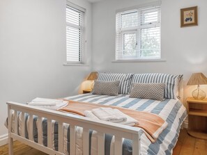 Quarto com cama, almofadas, mesa de cabeceira, abajur e duas janelas com persianas.