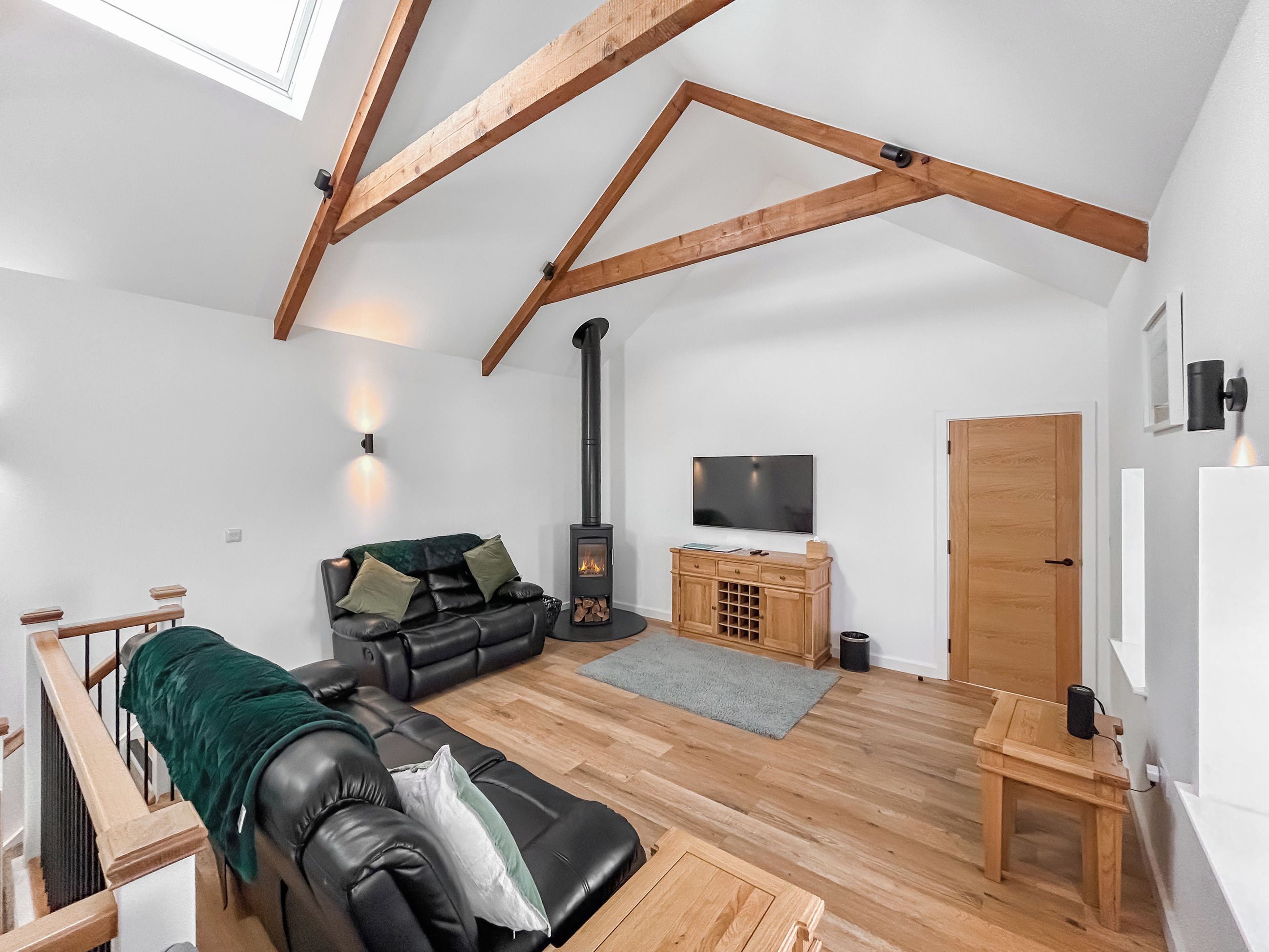A loft-style living room with exposed wooden beams, a black leather sofa, a television, a wooden cabinet, and a fireplace.