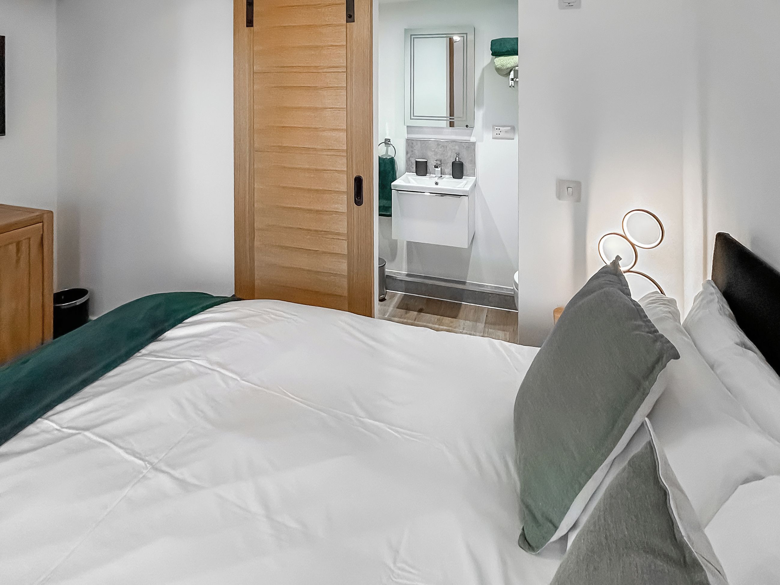 A bedroom with a bed, a wooden door, a bathroom with a sink and mirror, and a green towel.