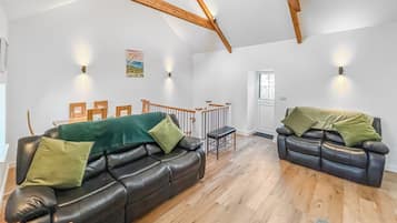 Amplia sala de estar con techo alto, vigas de madera, dos sofás de cuero con cojines verdes, una escalera de madera y una puerta que da al exterior.