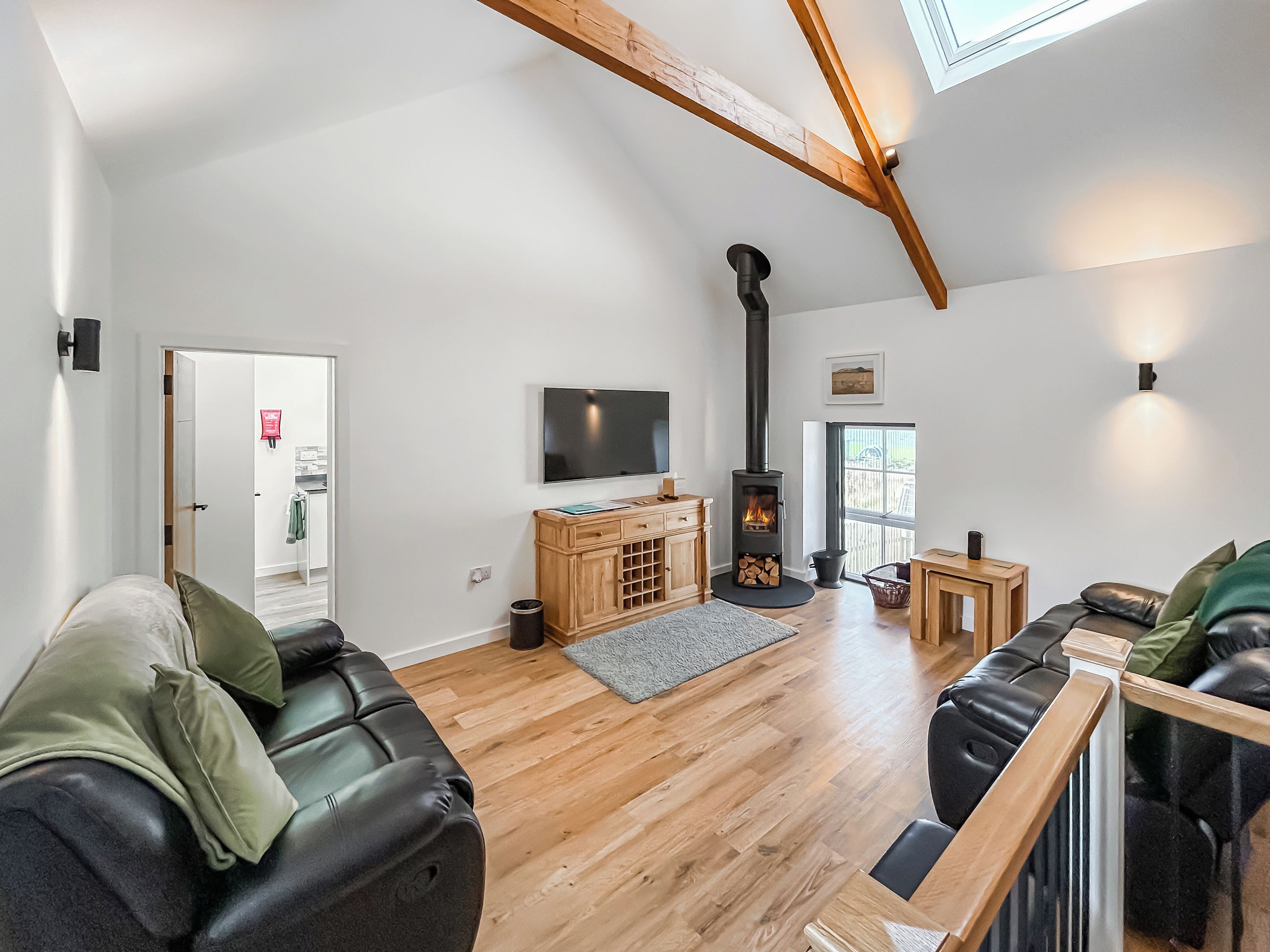 A modern living room with a wooden floor, a black leather sofa, a TV mounted on the wall, and a wood-burning stove.