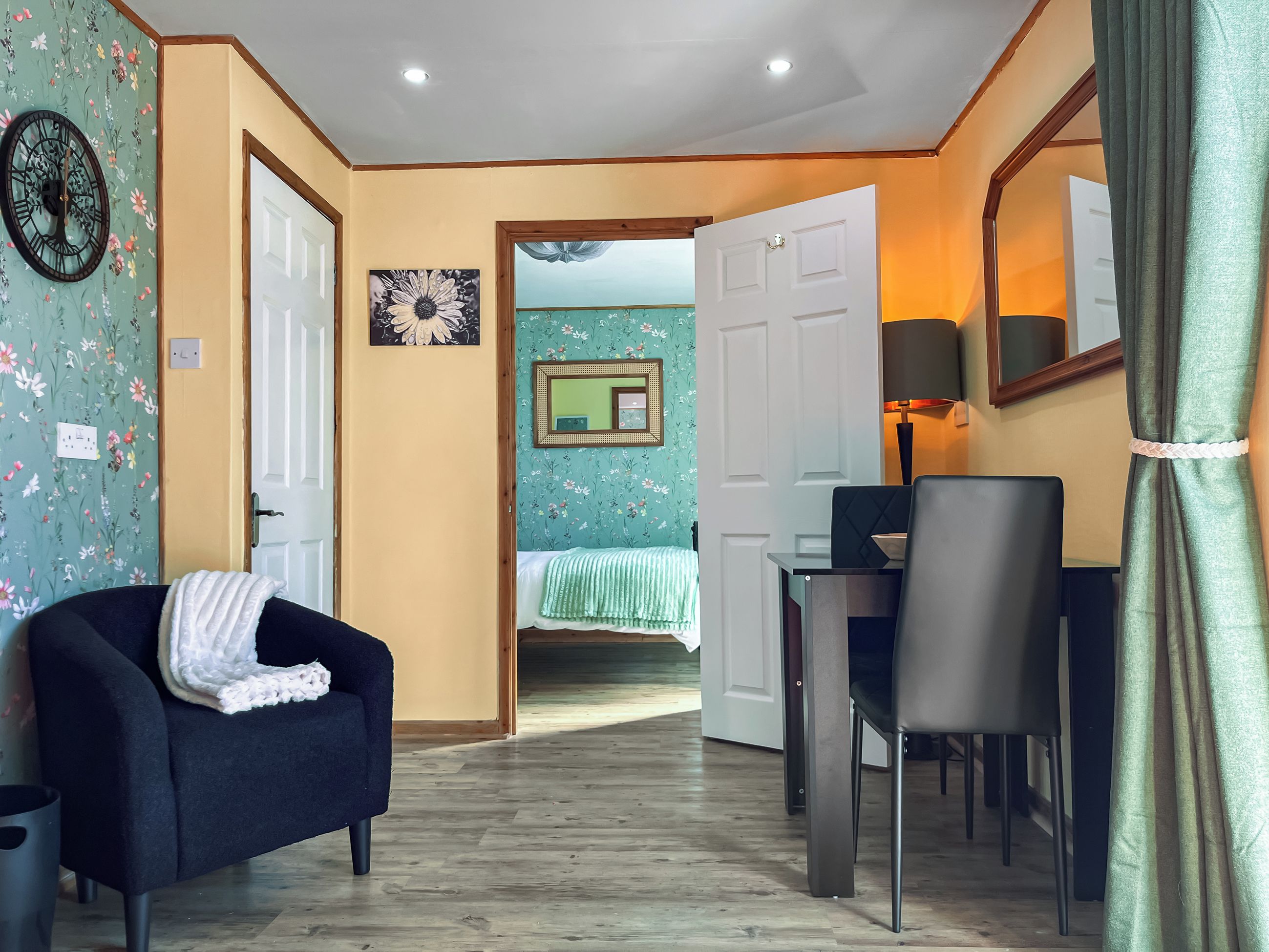 Una habitación con sofá, mesa de comedor con sillas, cama con colcha verde, un reloj de pared y un espejo.
