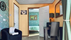 Una habitación con sofá, mesa de comedor con sillas, cama con colcha verde, un reloj de pared y un espejo.