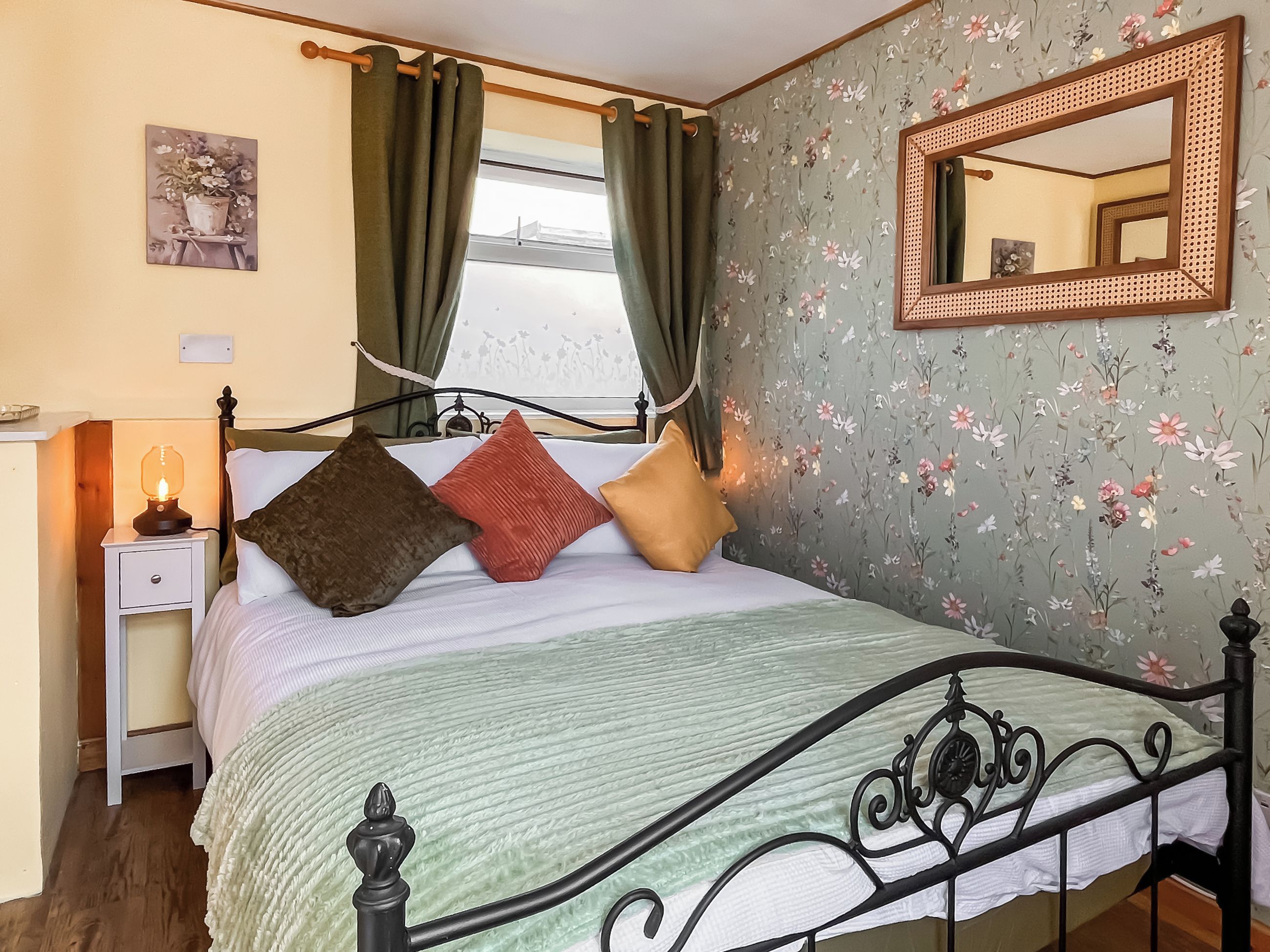 Ein Bett mit Kopfteil, ein Nachttisch mit Lampe und blumigem Wandpapier.