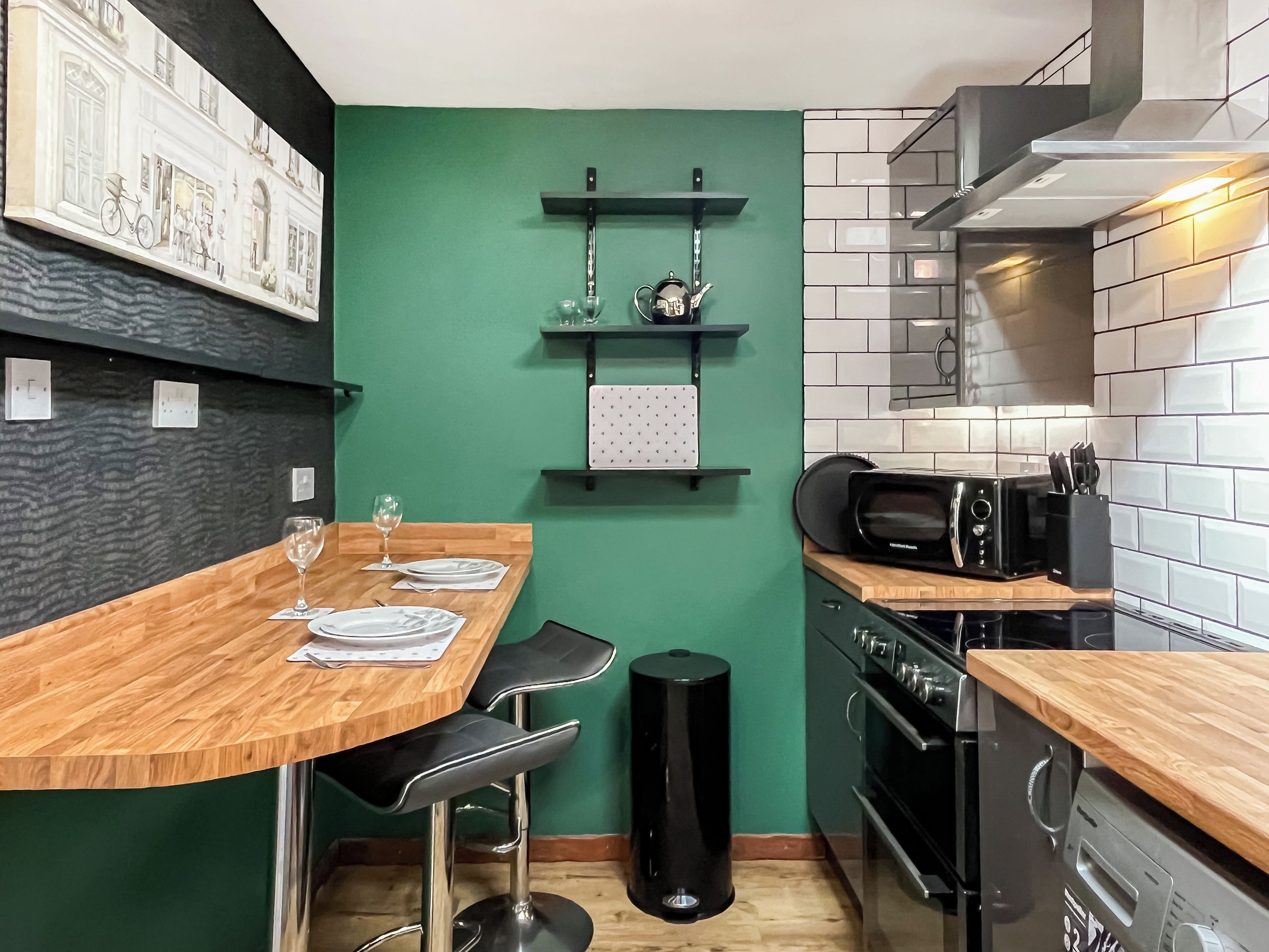 Una cocina moderna con una pared de acento verde, una mesa de comedor de madera, taburetes negros y un cubo de basura negro.