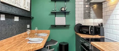 A modern kitchen with a green accent wall, a wooden dining table, black bar stools, and a black trash bin.