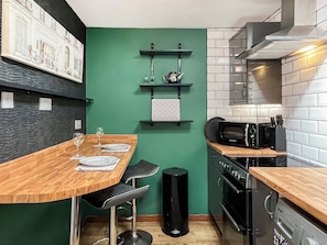 A modern kitchen with a green accent wall, a wooden dining table, black bar stools, and a black trash bin.