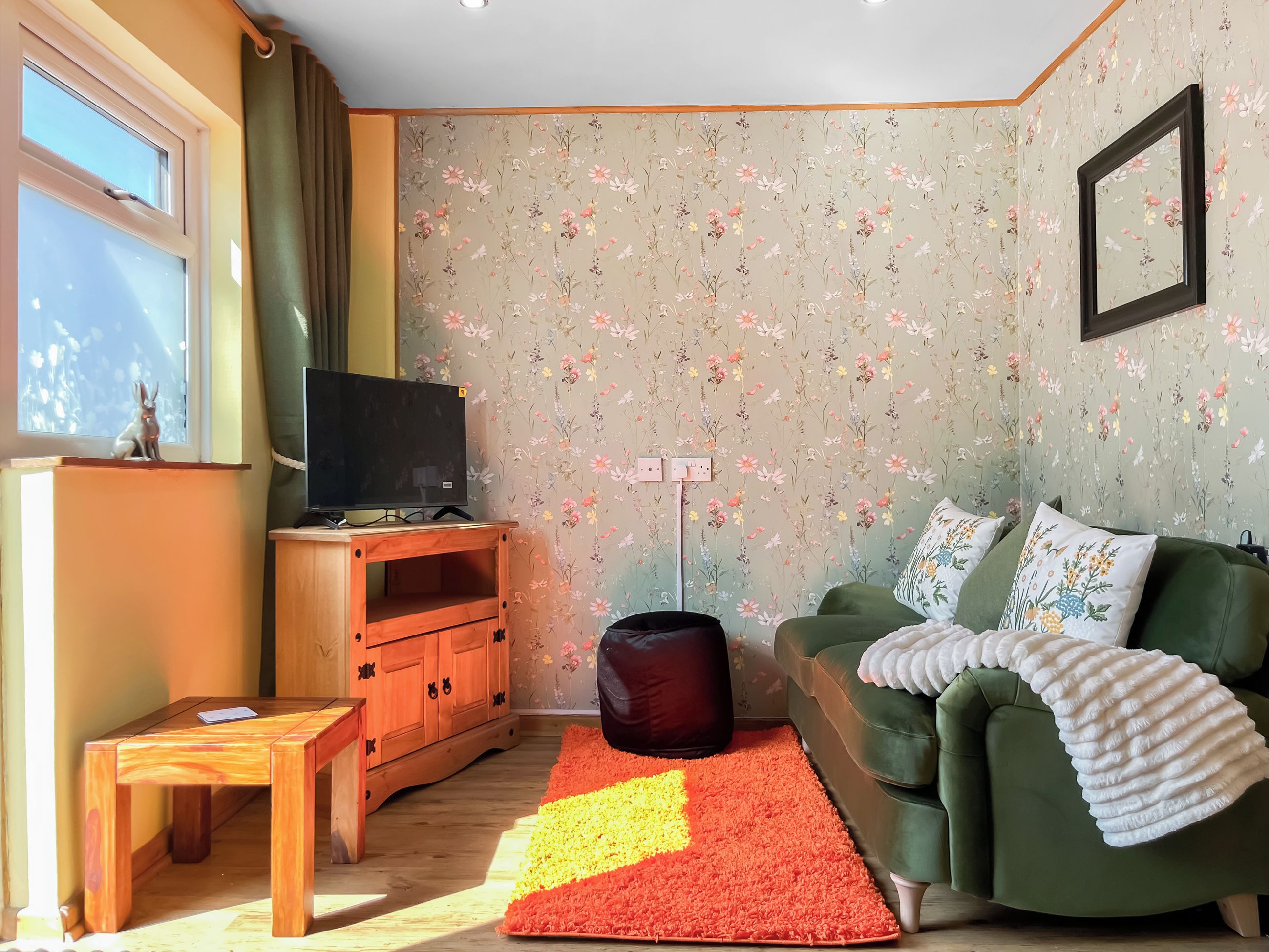 Una sala de estar con un televisor sobre un soporte de madera, un sofá verde con cojines florales, un tapete rojo y amarillo, y un papel tapiz floral.
