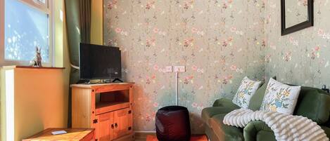 A living room with a television on a wooden stand, a green sofa with floral cushions, a red and yellow rug, and a floral wallpaper.