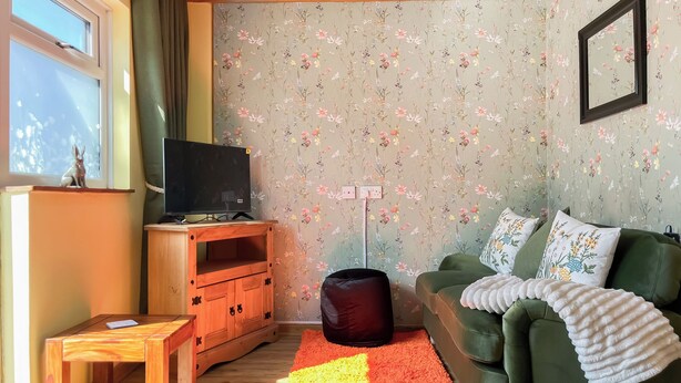 Una sala de estar con un televisor sobre un soporte de madera, un sofá verde con cojines florales, un tapete rojo y amarillo, y un papel tapiz floral.