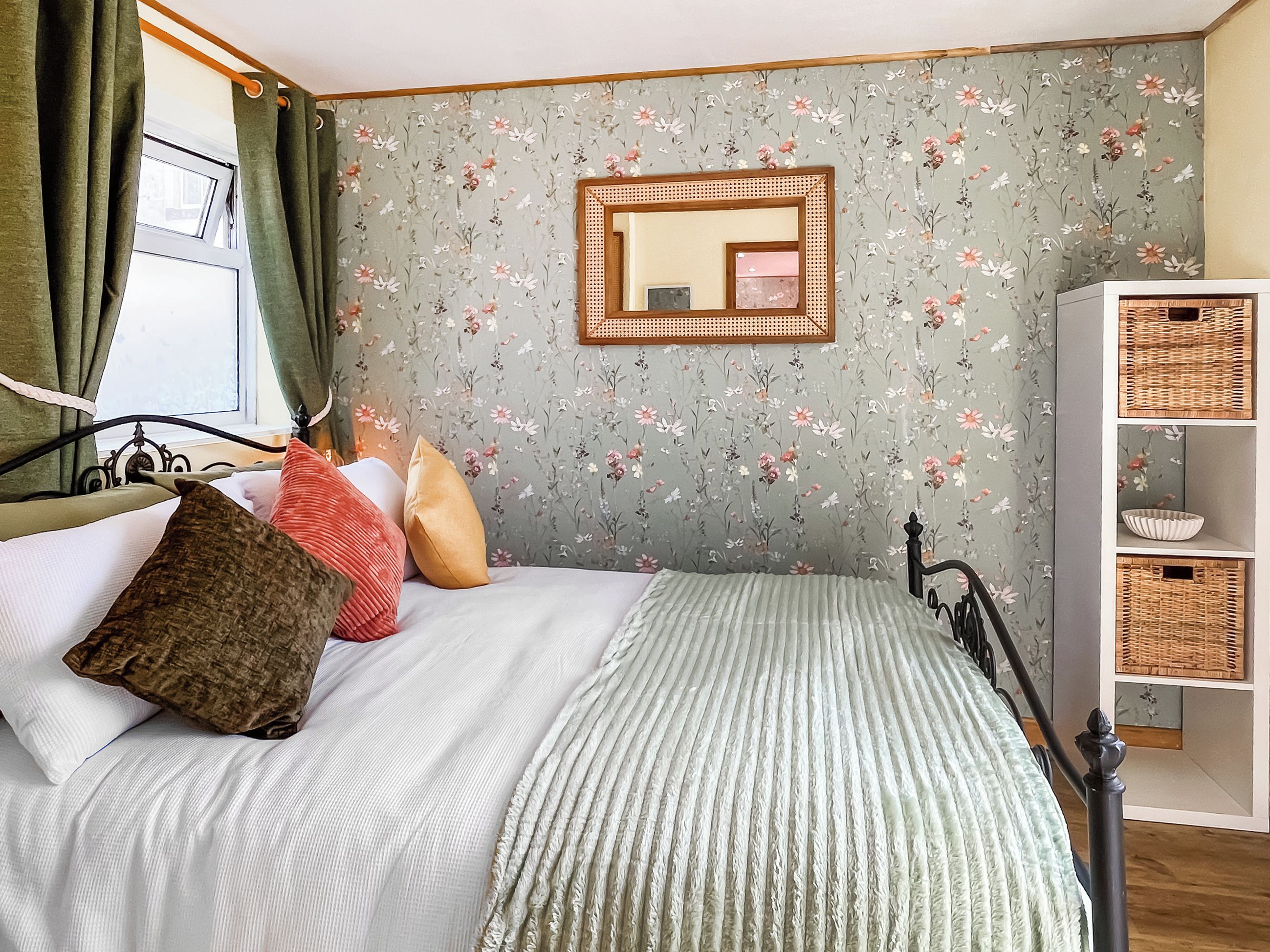 A bedroom with a bed, pillows, a bedside table, a mirror, and a wall with floral wallpaper.