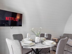Uma área de refeições com uma mesa redonda posta para quatro pessoas, uma televisão na parede exibindo 'NETFLIX' e um sofá cinza ao fundo.