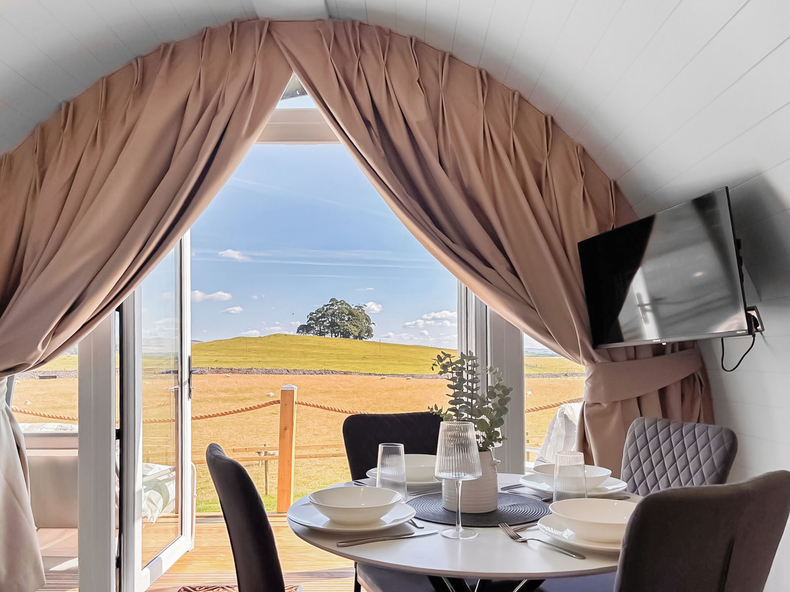 Ein Essbereich mit einem für zwei Personen gedecktem Tisch, einem an der Wand montierten Fernseher und Blick auf eine grüne Landschaft durch große Fenster.