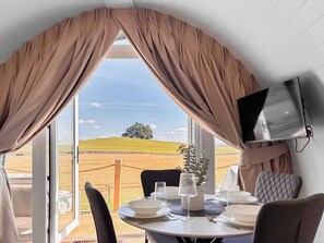 Ein Essbereich mit einem für zwei Personen gedecktem Tisch, einem an der Wand montierten Fernseher und Blick auf eine grüne Landschaft durch große Fenster.