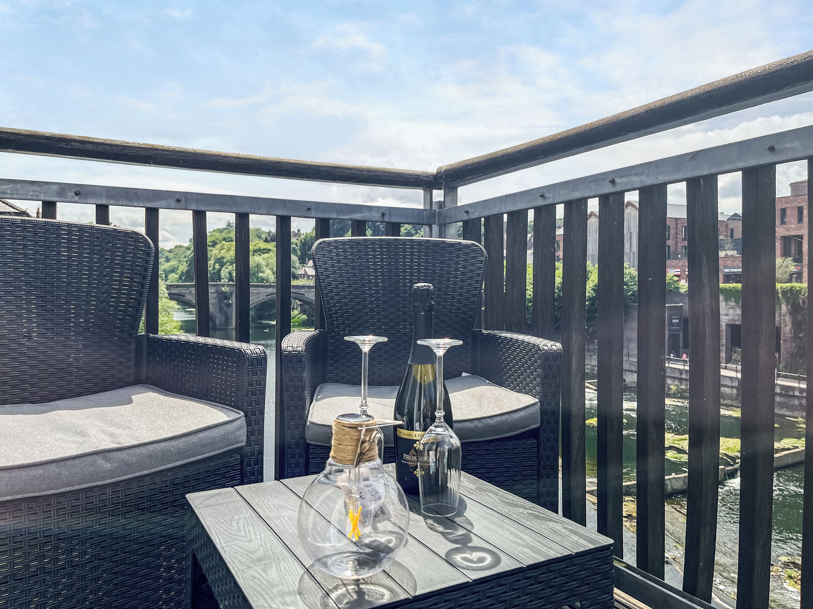 Un balcon agrémenté de meubles en osier, une table avec une bouteille et un verre, et une vue sur une rivière et des bâtiments.