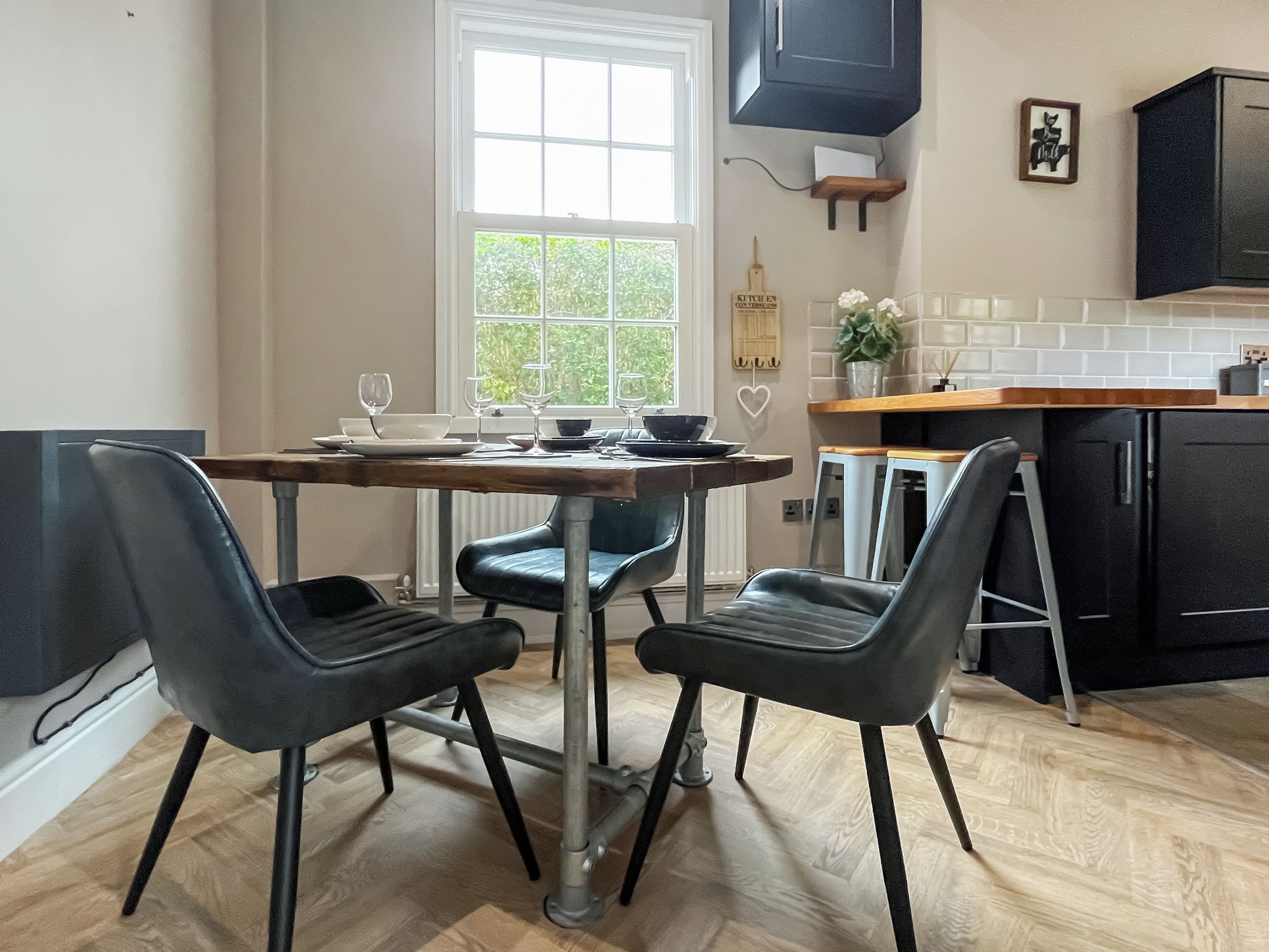 A modern kitchen with a dining table set for four, two black chairs, and a window with a view of greenery.