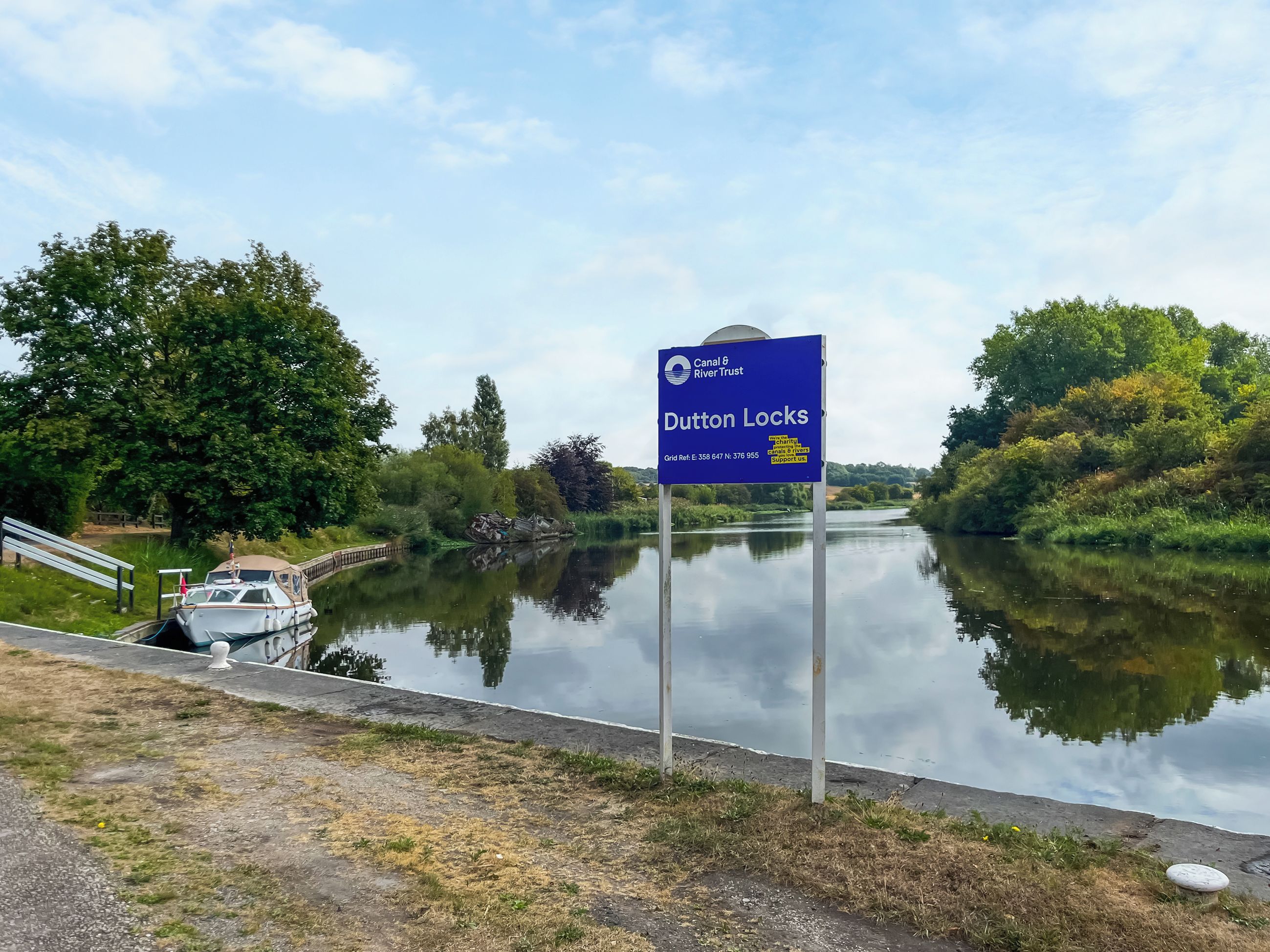 Un canal con un cartel que indica Dutton Locks, una embarcación amarrada a la izquierda y una zona ajardinada con un banco.