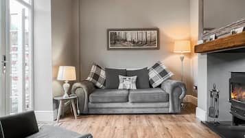 A living room with a grey sofa, a small side table with a lamp, a fireplace, and a large window.