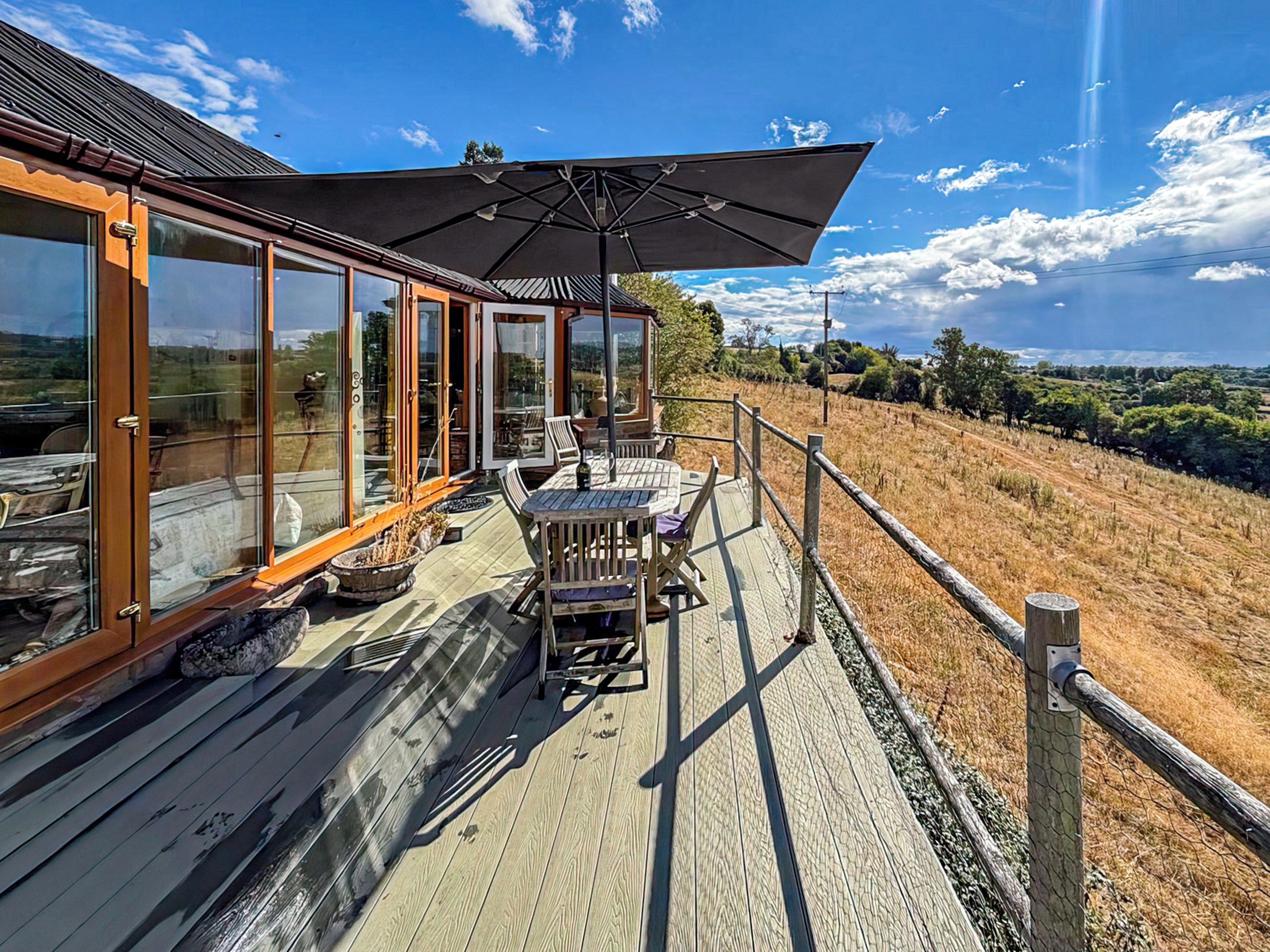A wooden deck with a table and chairs, a large umbrella, and a glass-enclosed room with doors.