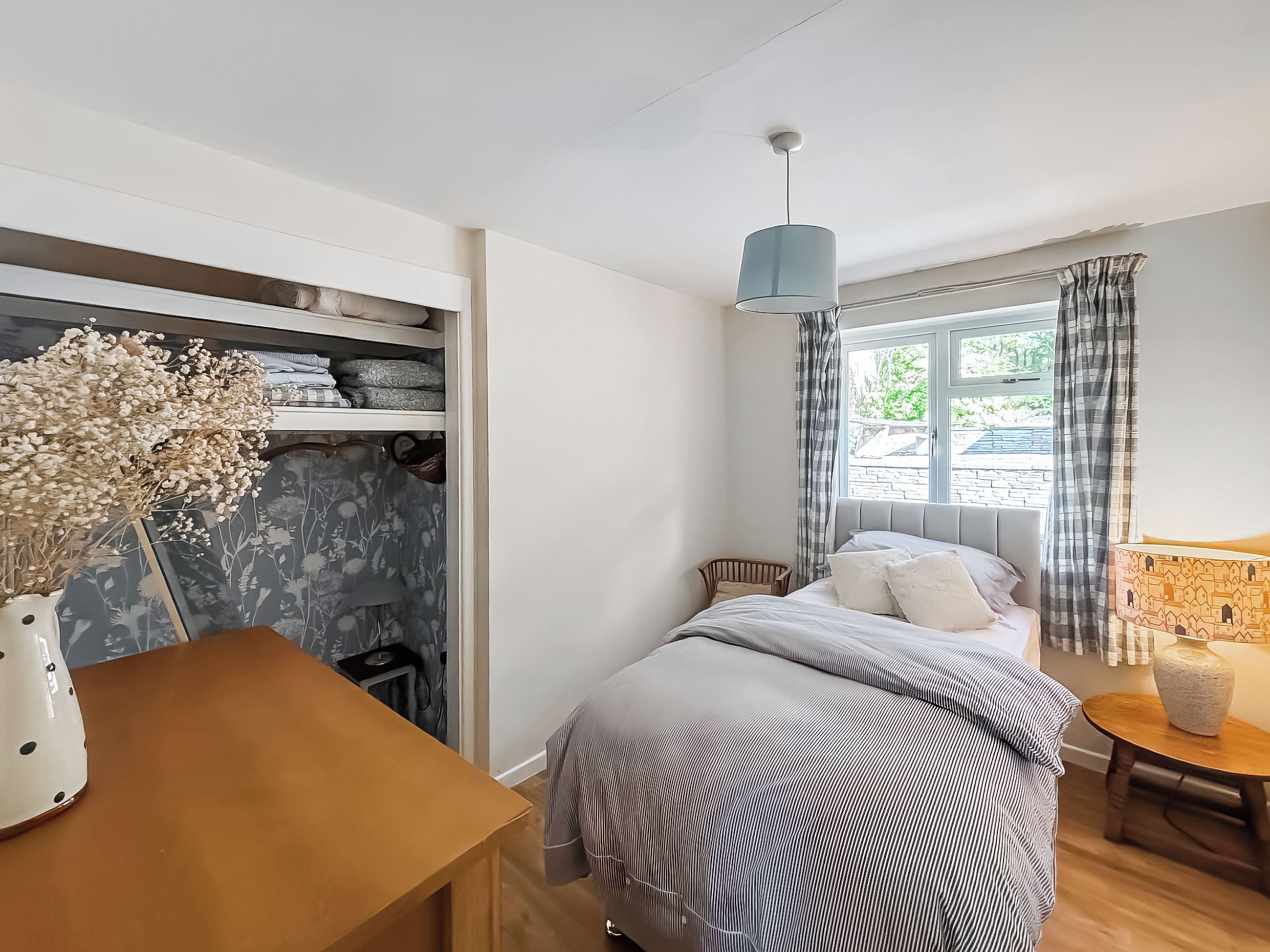 A bedroom with a bed, a bedside table, a lamp, a window with curtains, and a closet with shelves.