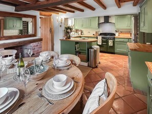 A dining area with a wooden table set for a meal, a kitchen in the background with green cabinets, and a brick wall. - 4 bedroom accommodation in Old Dalby (Old Dalby)