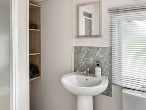 Een badkamer met een witte wastafel, een spiegel en een toilet. Er is een houten plankje met handdoeken en een zeepfles aan de muur.