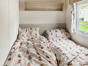 Een enkel bed met een geruite sprei met teddyberen erop, een houten hoofdbord en een raam met uitzicht op een huis.