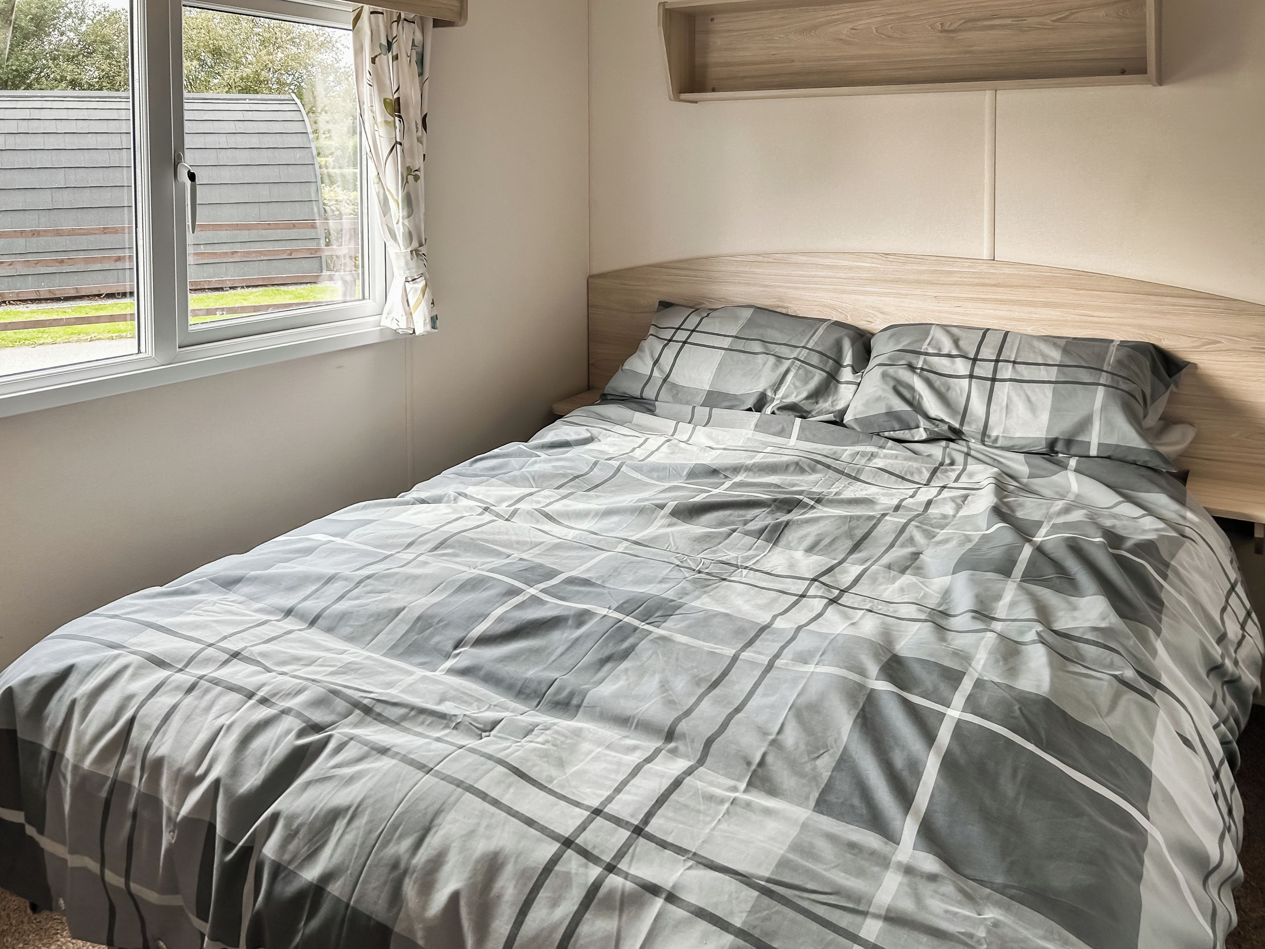 Una cama con colcha de cuadros grises y blancos, cabecera de madera y una ventana con cortinas.