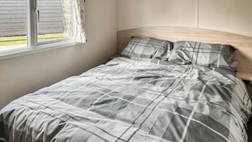 Una cama con colcha de cuadros grises y blancos, cabecera de madera y una ventana con cortinas.
