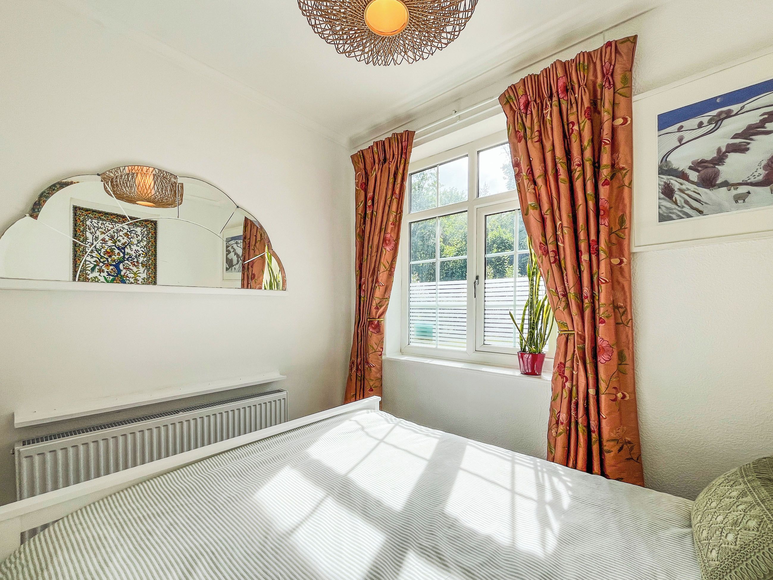 A bedroom with a bed, a window with curtains, a radiator, a potted plant, and a wall-mounted mirror.