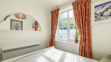 A bedroom with a bed, a window with curtains, a radiator, a potted plant, and a wall-mounted mirror.
