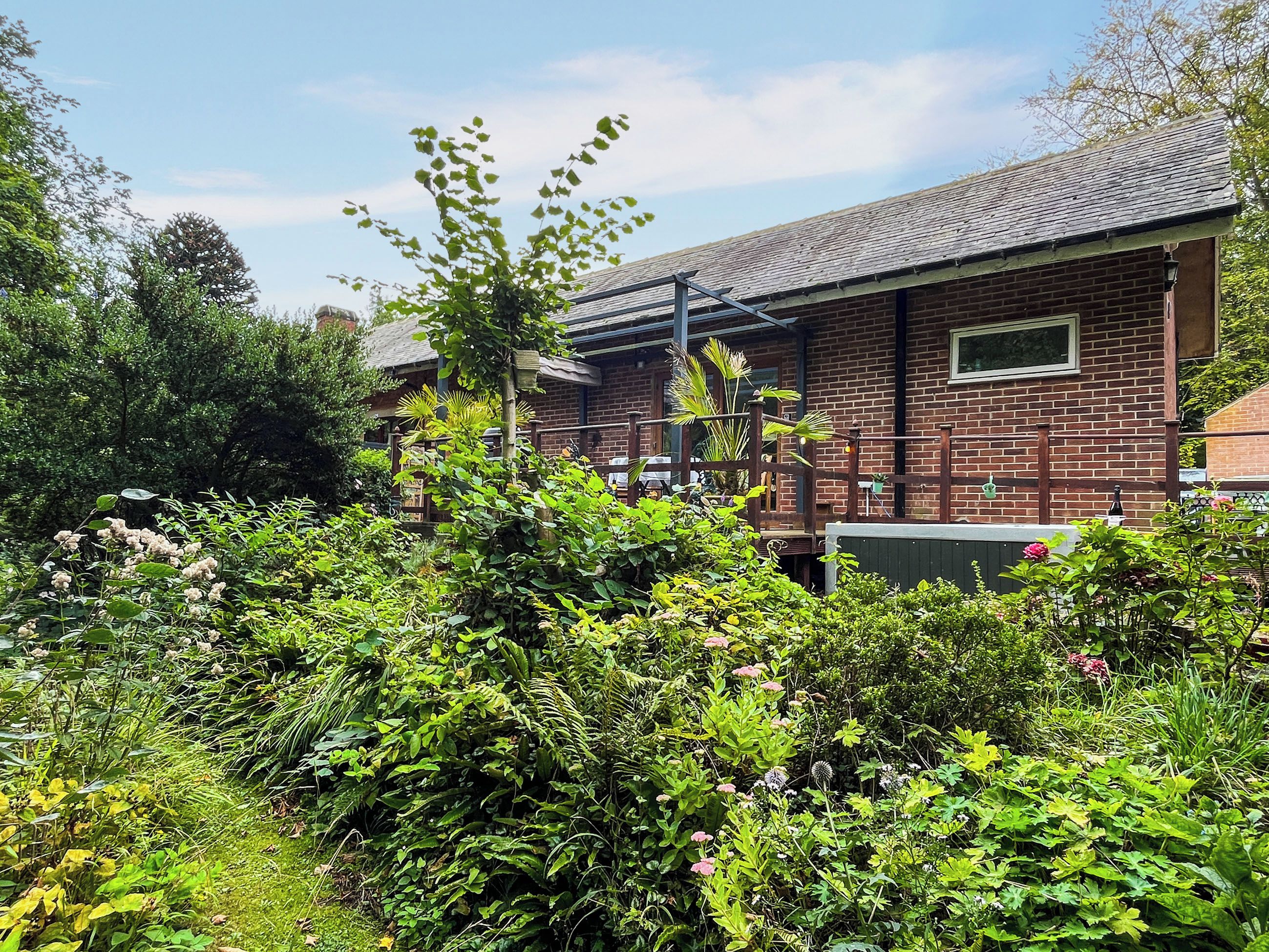 A brick building with a metal roof, surrounded by dense greenery and flowering plants.