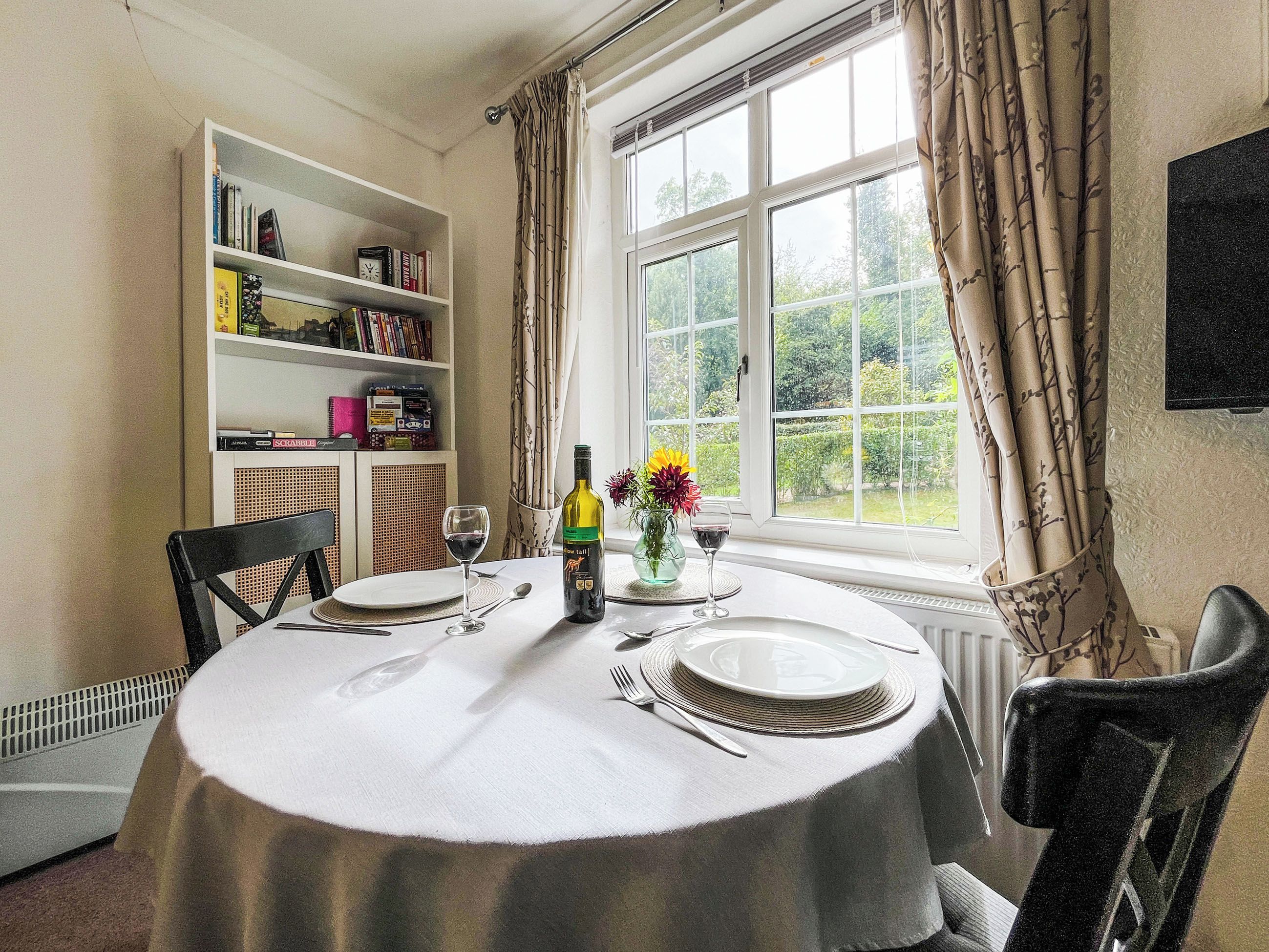 A dining table set with plates, cutlery, and wine glasses, with a bookshelf and curtains in the background.