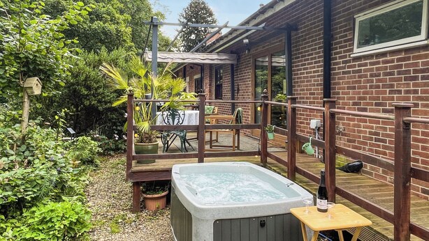 Ein Whirlpool in einem Garten mit einer Holzterrasse, umgeben von viel Grün und einem Backsteinhaus.