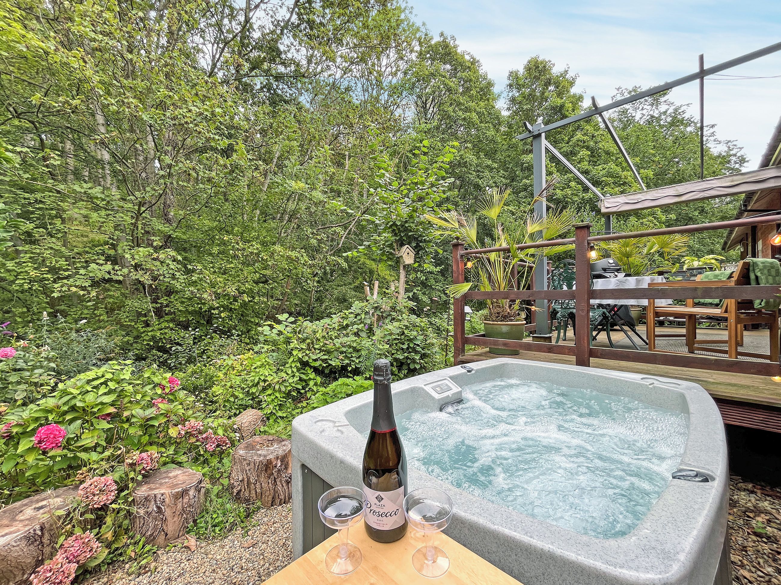 A hot tub with a bottle of wine and two glasses on a wooden table, surrounded by lush greenery and a wooden deck.