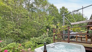 Ein Whirlpool mit einer Weinflasche und zwei Gläsern auf einem Holztisch, umgeben von üppigem Grün und einer Holzterrasse.